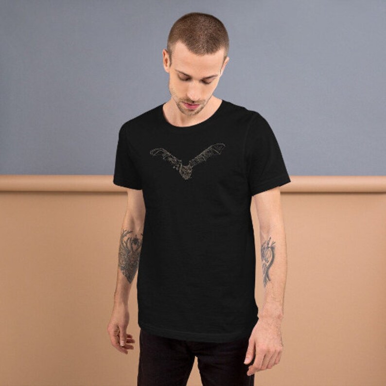 K&ouml;nnte beinhalten: Schwarzes T-Shirt mit einem goldfarbenen Fledermaus-Design auf der Brust. Die Fledermaus ist mit ausgebreiteten Fl&uuml;geln dargestellt. Das T-Shirt besteht aus einem weichen Material und wird von einer Person getragen.