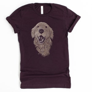 May include: A dark purple t-shirt with a detailed illustration of a golden retriever dog's head. The dog is smiling with its tongue out.