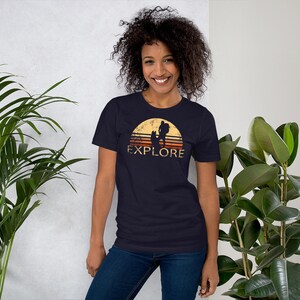 May include: Navy blue t-shirt featuring a retro design with a hiker and dog silhouette set against a sunset. The word "EXPLORE" is printed below the graphic in a vintage font. The shirt is made of a soft material.