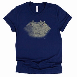May include: Navy blue t-shirt featuring a detailed illustration of a frog's face. The frog's face is in shades of gray and tan, with orange eyes. The shirt has rolled-up sleeves and a tag on the upper back.
