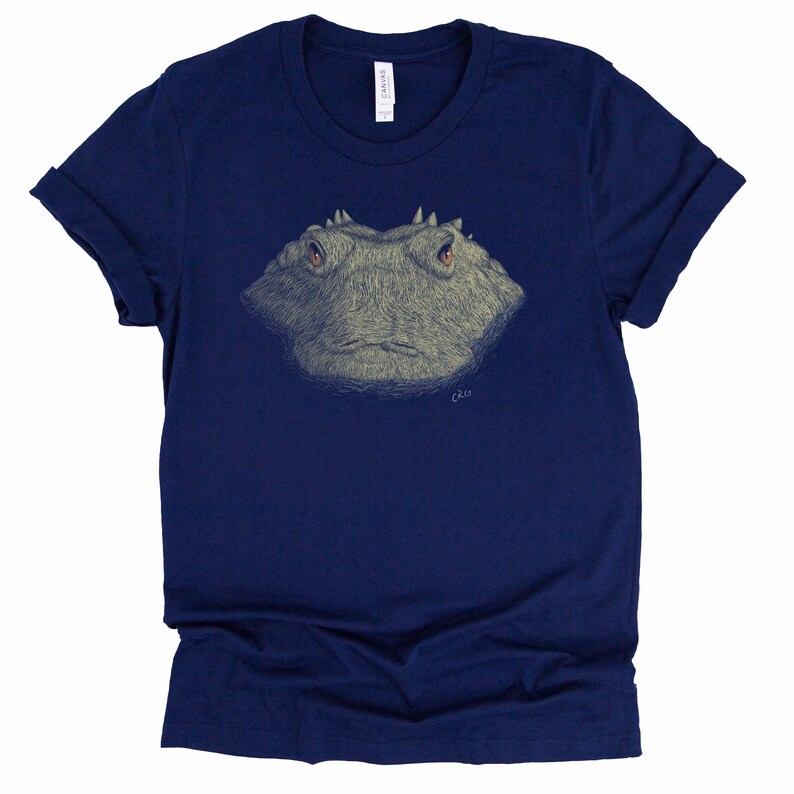 May include: Navy blue t-shirt featuring a detailed illustration of a frog's face. The frog's face is in shades of gray and tan, with orange eyes. The shirt has rolled-up sleeves and a tag on the upper back.