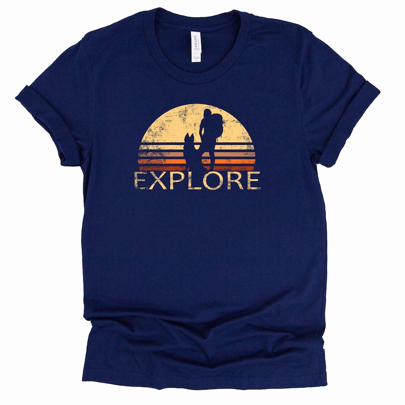 May include: Navy blue t-shirt with a distressed graphic of a hiker with a dog silhouetted against a sunset. The word "EXPLORE" is printed below the image in a vintage font. The shirt is made of soft material.