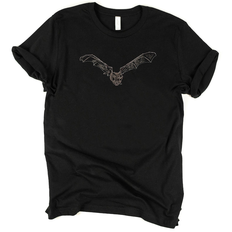 K&ouml;nnte beinhalten: Schwarzes T-Shirt mit einer detaillierten Illustration einer Fledermaus im Flug. Die Fledermaus ist mit ausgebreiteten Fl&uuml;geln dargestellt, die filigrane Linienarbeit zeigen. Das Design ist auf dem T-Shirt zentriert und bietet einen auff&auml;lligen Anblick auf dem dunklen Stoff. Das T-Shirt hat hochgekrempelte &Auml;rmel.