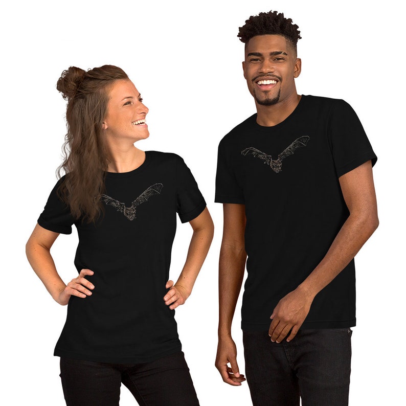 K&ouml;nnte beinhalten: Zwei Personen tragen schwarze T-Shirts. Jedes Shirt hat eine detaillierte, stilisierte Illustration einer Fledermaus mit ausgebreiteten Fl&uuml;geln. Das Fledermaus-Design ist auf der Brust zentriert.