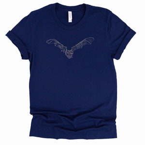 K&ouml;nnte beinhalten: Marineblaues T-Shirt mit einer detaillierten, hellfarbigen Illustration einer Fledermaus im Flug. Die Fl&uuml;gel der Fledermaus sind weit ausgebreitet, und das Design ist auf dem Shirt zentriert. Das Shirt hat hochgekrempelte &Auml;rmel und einen klassischen Rundhalsausschnitt.