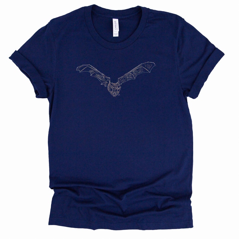 K&ouml;nnte beinhalten: Marineblaues T-Shirt mit einer detaillierten, hellfarbigen Illustration einer Fledermaus im Flug. Die Fl&uuml;gel der Fledermaus sind weit ausgebreitet, und das Design ist auf dem Shirt zentriert. Das Shirt hat hochgekrempelte &Auml;rmel und einen klassischen Rundhalsausschnitt.