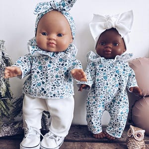 Peut inclure: Deux poupées pour bébés vêtues de tenues fleuries assorties. L'une porte un bandeau et un pantalon, l'autre une barboteuse et un nœud blanc. Les deux poupées ont un col à volants et des chaussures blanches.