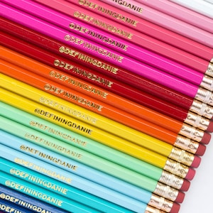 May include: A diagonal arrangement of coloured pencils, spanning a rainbow of shades from blue to pink. Each pencil features a gold ferrule and eraser, with the text "@DEFININGDANIE" printed in gold.