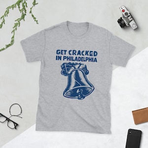 Könnte beinhalten: Ein kurzärmeliges T-Shirt in Grau meliert mit einer dunkelblauen Grafik der Liberty Bell und dem Text "GET CRACKED IN PHILADELPHIA", der über der Glockenillustration gedruckt ist. Das Design ist mittig auf der Brust des Kleidungsstücks platziert.