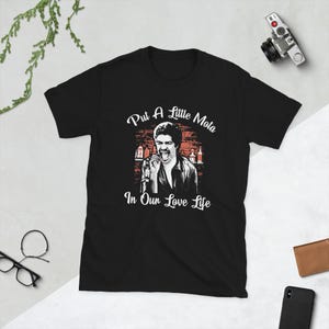 May include: Black t-shirt featuring a graphic of a man holding a microphone with the text "Put A Little Mota In Our Love Life". The design includes a bar scene with bottles. A vintage camera is visible in the background.