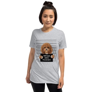 May include: Light gray t-shirt featuring a brown dog with a mugshot background. The dog holds a sign that reads "BAD DOG" with the numbers "230570" and "POLICE DEPT." printed below. The shirt has a crew neck and short sleeves.