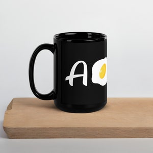 May include: Black ceramic mug with a white graphic of a fried egg and the letter "A".