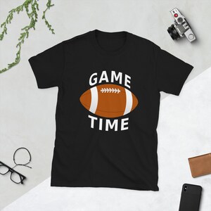 May include: Black t-shirt with the words "GAME TIME" in white, above and below a brown and white football graphic. The shirt is a short-sleeved crew neck.