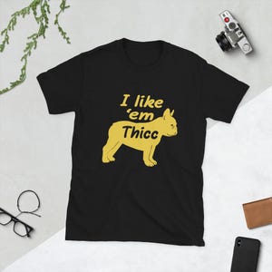 May include: Black t-shirt with a yellow graphic of a French Bulldog and the text "I like 'em Thicc". The shirt is made of a soft material and has short sleeves. The design is centered on the front of the shirt.