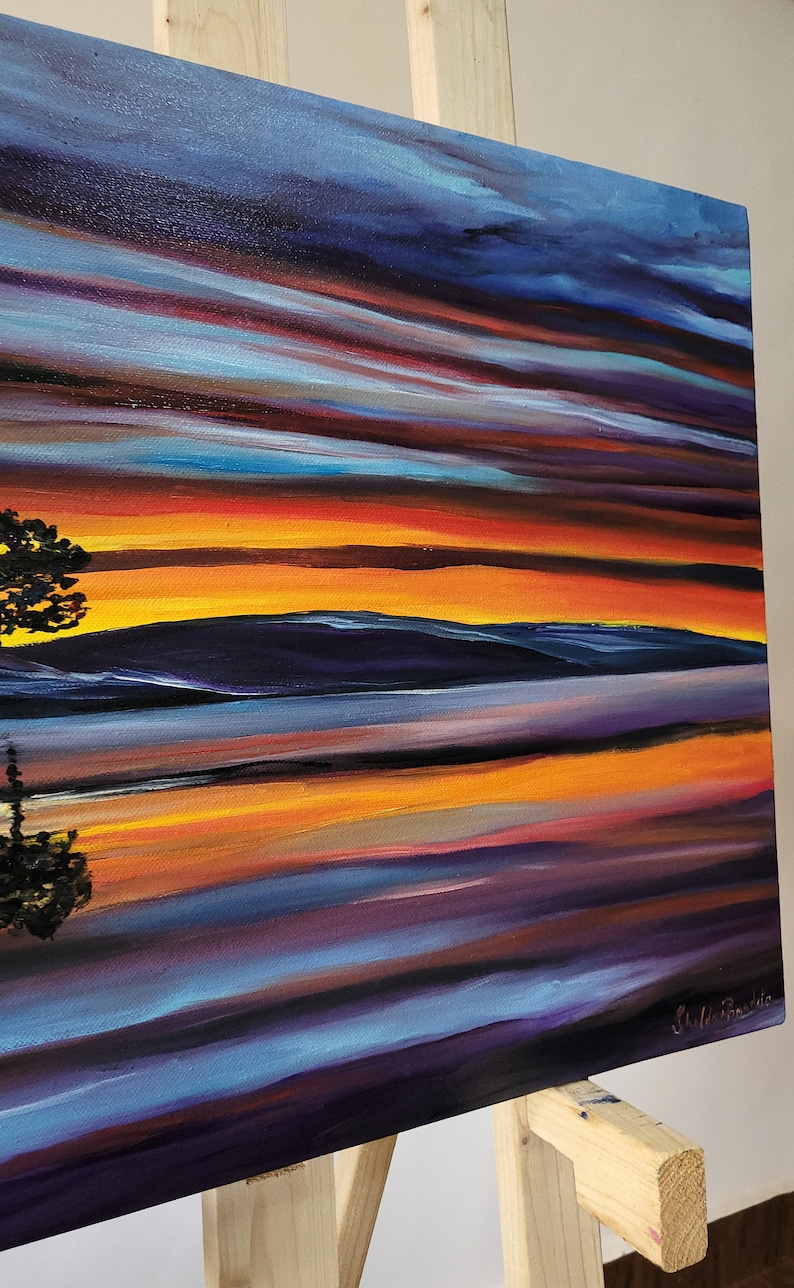 May include: An original oil painting on canvas depicting a vibrant sunset over a body of water. The artwork features bold brushstrokes in shades of orange, yellow, blue, and purple, with a silhouette of a tree in the foreground. The painting is displayed on a wooden easel.