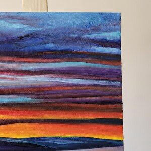May include: A close-up of a canvas painting depicting a sunset. The artwork features horizontal bands of color, including shades of blue, purple, orange, and yellow. The painting is mounted on a wooden easel.