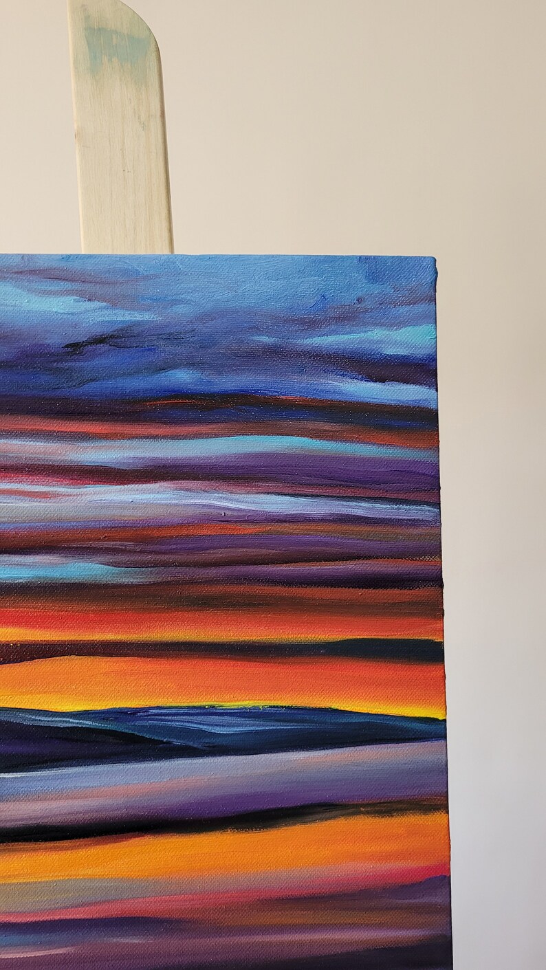 May include: A close-up of a canvas painting depicting a sunset. The artwork features horizontal bands of color, including shades of blue, purple, orange, and yellow. The painting is mounted on a wooden easel.