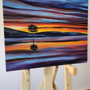 May include: An original oil painting on canvas depicting a sunset scene with a tree silhouette reflected in the water. The artwork features vibrant colors of orange, purple, and blue. The painting is displayed on a wooden easel.