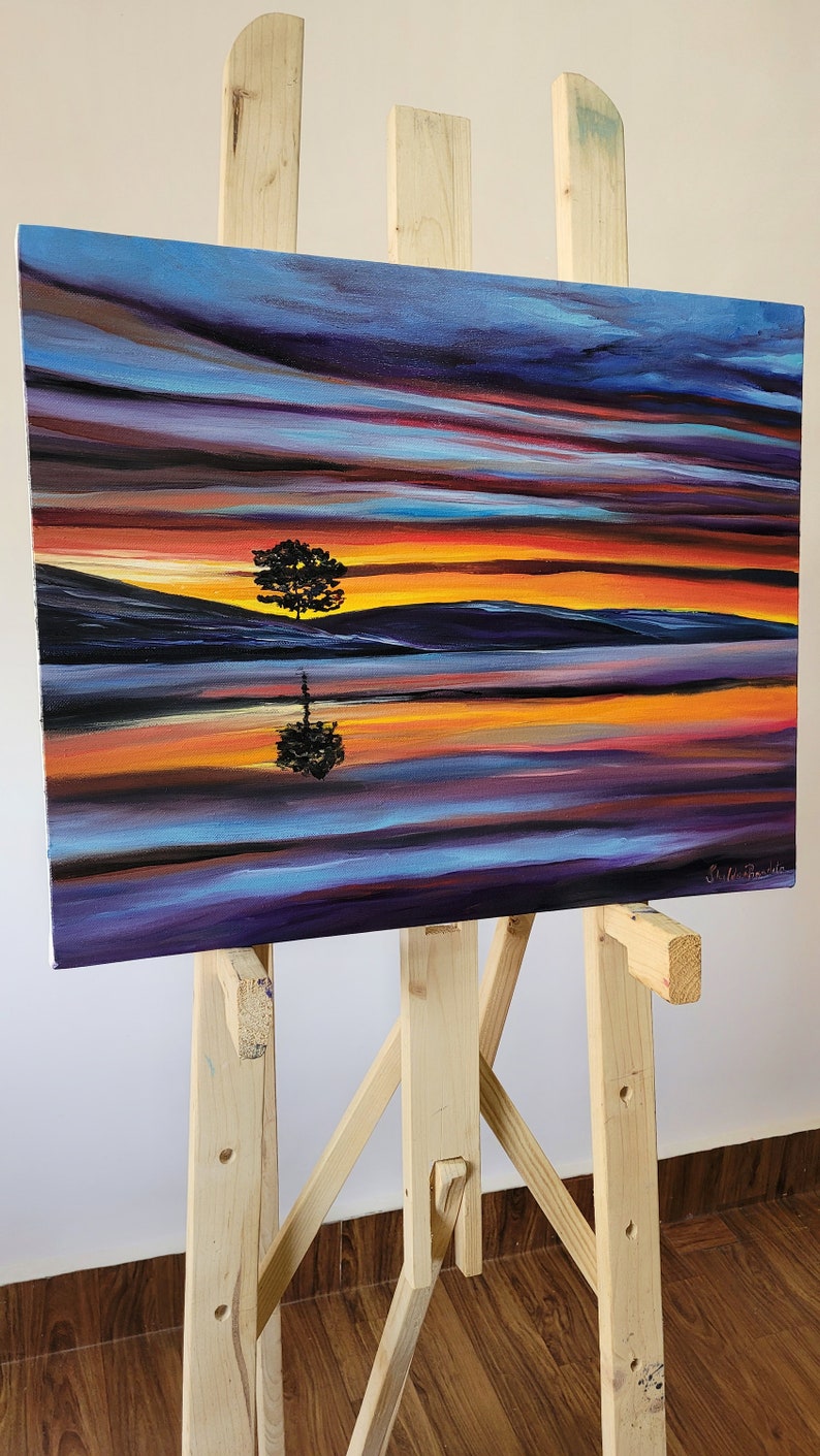 May include: An original oil painting on canvas depicting a sunset scene with a tree silhouette reflected in the water. The artwork features vibrant colors of orange, purple, and blue. The painting is displayed on a wooden easel.