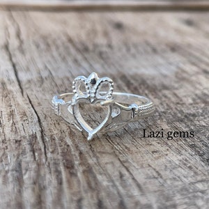May include: A silver Claddagh ring with a heart, crown, and hands design. The ring is on a wooden surface.