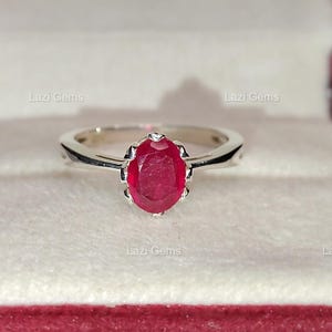 Peut inclure: Une bague en argent ornée d'une pierre précieuse rouge ovale. La bague est sertie dans un style classique, mettant en valeur la couleur riche du rubis. La bague est présentée dans un écrin rouge et blanc.