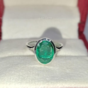 Puede incluir: Un anillo de plata con una gran esmeralda de talla ovalada. La piedra verde brillante está engastada en una banda sencilla y elegante. El anillo se muestra en una caja de joyería roja con interior blanco. El anillo es una joya.