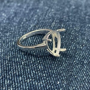 Pode incluir: Uma base de anel de prata com um design em forma de pêra. O anel é feito de um material metálico brilhante e apresenta garras para segurar uma pedra preciosa. O anel está sobre um fundo de ganga azul.