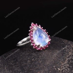 May include: A pear-shaped ring featuring a large, iridescent moonstone center, surrounded by small, vibrant red gemstones. The ring has a silver band and is set against a dark, textured background. The ring is a piece of jewelry.
