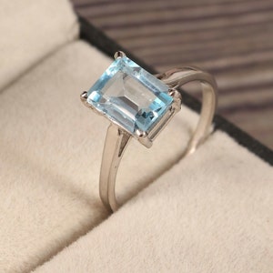 Puede incluir: Anillo de plata con una piedra preciosa rectangular de color azul claro. La piedra está engastada en un engaste de cuatro garras, y la banda es de plata pulida. El anillo se muestra en una caja de joyería.