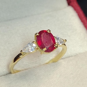 May include: A gold ring featuring a large oval red ruby gemstone and two smaller pear-shaped clear gemstones.