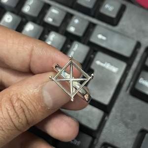 Könnte beinhalten: Ein silberner Ring-Einsatz mit einem quadratischen, offenen Zentrum, der für einen Edelstein ausgelegt ist. Der Ring wird an einem Finger gezeigt.