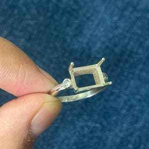 May include: A silver-toned ring setting featuring a rectangular prong head designed to hold a center stone. The band includes a small, round, clear accent stone on each side of the central mounting, providing a decorative element to the metal ring.