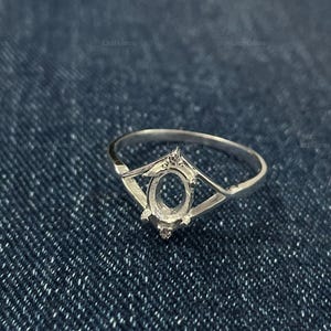 Pode incluir: Um anel de prata com um engaste oval, projetado para segurar uma pedra preciosa. O anel apresenta um design geométrico decorativo ao redor do engaste e uma banda delicada. O anel é exibido em uma superfície de ganga azul.