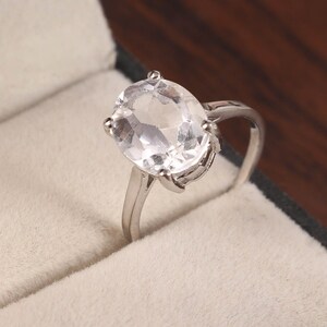 Può includere: Un anello d'argento con una grande gemma trasparente a taglio ovale. La gemma è tenuta in posizione da quattro griffe. L'anello è posto su uno sfondo bianco sporco, forse un portagioie.