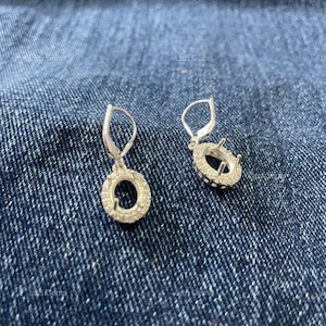 May include: A pair of silver dangle earrings with a circular design. The earrings feature a halo of small, clear gemstones around the outer edge of the circle and are set against a blue denim background.