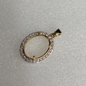 May include: A gold-colored pendant with an oval-shaped frame, encrusted with small, clear gemstones. The pendant has a loop at the top for a chain and is set against a light background. The text "Lazi Gems" is visible.