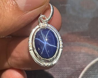 May include: A silver pendant featuring an oval, deep blue gemstone with a six-pointed star pattern. The gemstone is set in a silver bezel with decorative detailing. The pendant has a small loop for a chain.
