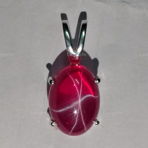 May include: A silver pendant featuring a red star ruby gemstone with white star-like inclusions. The pendant is oval shaped and has a silver bail.