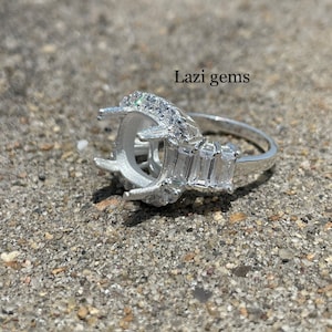 May include: A silver ring with a large oval setting surrounded by smaller stones. The ring also has baguette-shaped stones on the band. The ring is on a gray, pebbled surface. Lazi gems is written in the top right corner of the image.