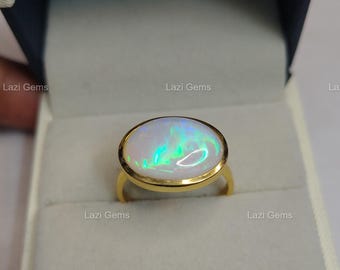 May include: A gold ring with an oval opal gemstone. The opal has a white base with iridescent flashes of green and blue. The ring is set in a gold band and is displayed in a white jewelry box.