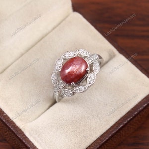May include: A silver ring with a red ruby gemstone surrounded by a halo of smaller diamonds. The ring is in a small, brown box.