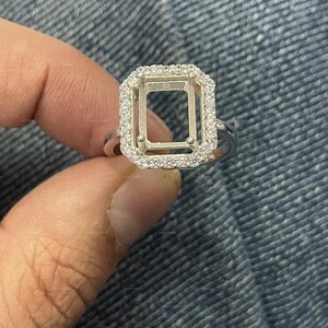 May include: A silver ring with a rectangular halo setting, encrusted with small, clear gemstones. The center is open, designed to hold a gemstone. The ring is held against a denim background.