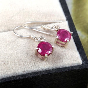 May include: A pair of silver earrings featuring red ruby gemstones. The earrings have a simple design with a round ruby stone set in a silver setting.