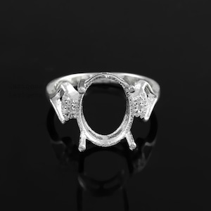 May include: A silver ring setting with an oval center and decorative accents. The ring features small, sparkling details on the sides and is designed to hold a gemstone. The ring is displayed on a reflective black surface.
