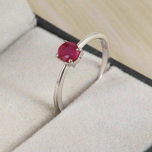 May include: A silver ring featuring a round, deep red gemstone. The gemstone is set in a four-prong setting, and the band is thin and polished. The ring is displayed in a gray jewelry box with a soft, white interior.