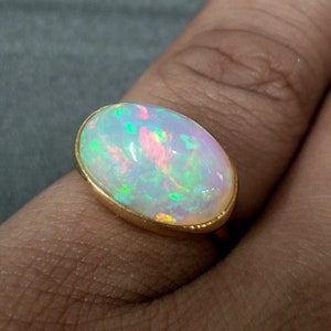 Op de afbeelding: Ovale opaalring met een gouden band. De opaal vertoont een kleurenspel met groene, roze en gele flitsen. De ring is gezet in een eenvoudige, klassieke gouden zetting, die de schoonheid van de edelsteen benadrukt.