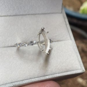 May include: A silver ring setting featuring a marquise-shaped prong mount with a textured finish. The band is adorned with small, clear, marquise-cut and round-cut stones set along the sides, creating a delicate and detailed appearance.