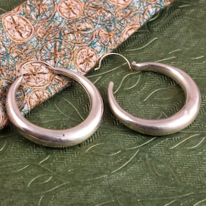 May include: A pair of silver hoop earrings with a smooth, rounded surface. The earrings are on a green patterned surface.