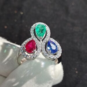 Peut inclure: Une bague en argent avec trois pierres précieuses en forme de poire : une émeraude verte, un rubis rouge et un saphir bleu. Les pierres précieuses sont serties dans un design délicat et complexe avec de petits diamants qui les entourent.