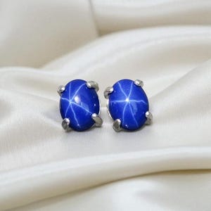 Pode incluir: Um par de brincos de botão ovais de safira estrela azul. Cada brinco apresenta uma gema azul vibrante com um padrão de estrela de seis raios, cravejada em metal prateado. Os brincos são exibidos em um tecido branco e sedoso.
